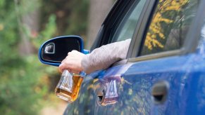 Driver holds a bottle of alcohol out the window of a blue car, trees visible in the background.