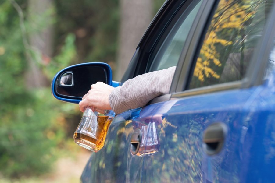 Driver holds a bottle of alcohol out the window of a blue car, trees visible in the background.