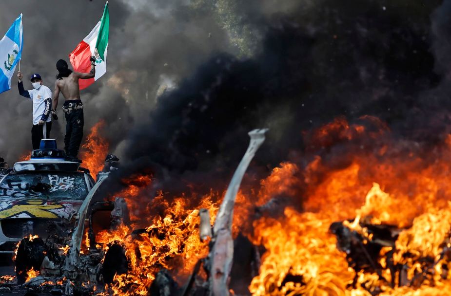 Protestors stand above burning Waymo taxi cars in downtown protests against ICE