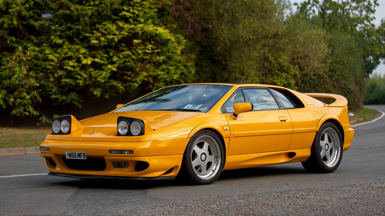Bright yellow 1990s Lotus Espirit supercar with pop-up headlights parked on a country road, trees visible in the background.