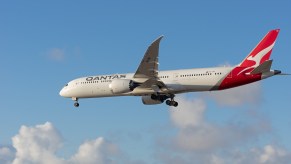 White Boeing plane with Qantas logo flies through a blue sky, clouds visible in the background.