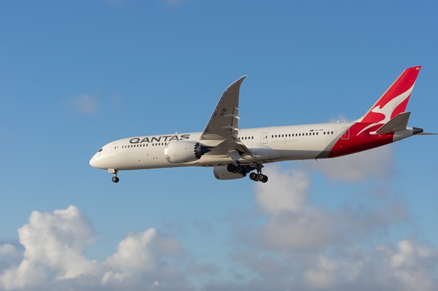 White Boeing plane with Qantas logo flies through a blue sky, clouds visible in the background.