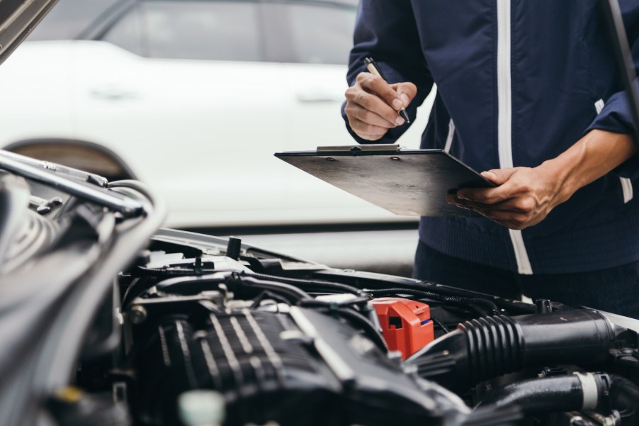 Mechanic completes a state-mandates safety inspection of a car, looking under its hood with a clipboard in his hand.
