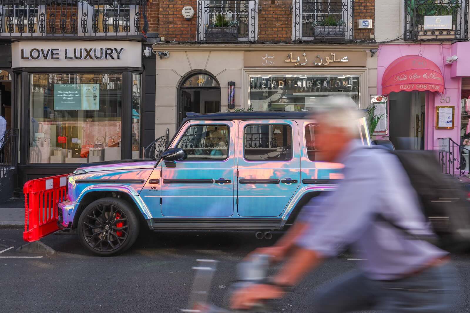 Used Mercedes G Wagon parked n the street by a luxury shop, a bicycler passes by in the foreground.