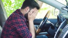 A man covers his face while sitting in the driver's seat of a car depicting frustration over car insurance costs