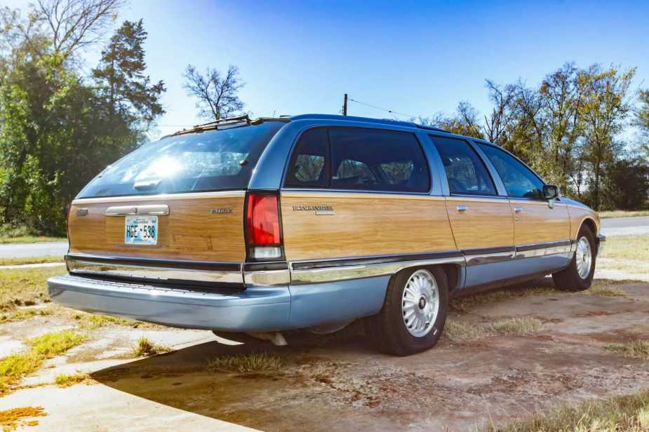 Rear view of a light blue Buick Roadmaster Estate Wagon with wood paneling, parked in front of a road with trees visible in the background.
