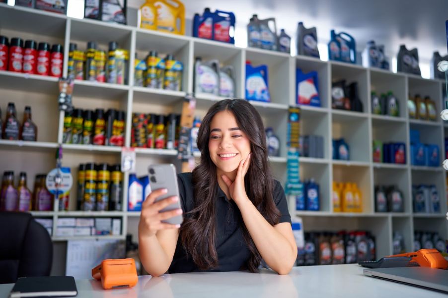 Saleswoman in an auto parts store, a shelf of A/C freon refrigerant refill canisters behind her.