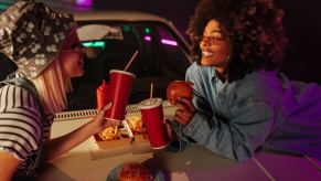 Two girls eat gas station food on the hood of their car.