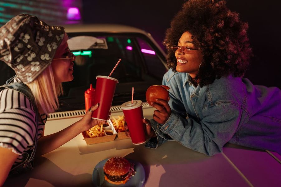Two girls eat gas station food on the hood of their car.
