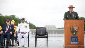 National Park Service speakers stands behind a podium next to military veterans to honor WWII