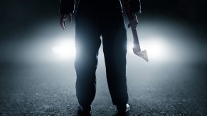 A man standing in front of a car's headlights at night holding a hatchet