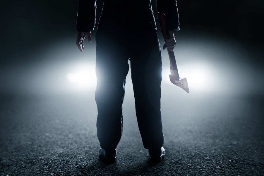 A man standing in front of a car's headlights at night holding a hatchet