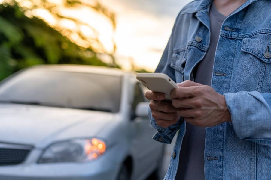 Man standing looking at his phone next to a car at dusk