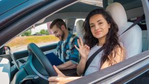 A new driver gestures at the camera while sitting behind the wheel.