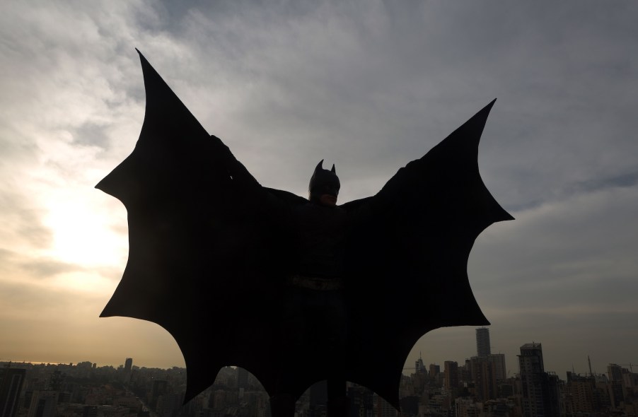 Man wearing Batman costume holds up his cape on a rooftop, a city skyline visible in the background.