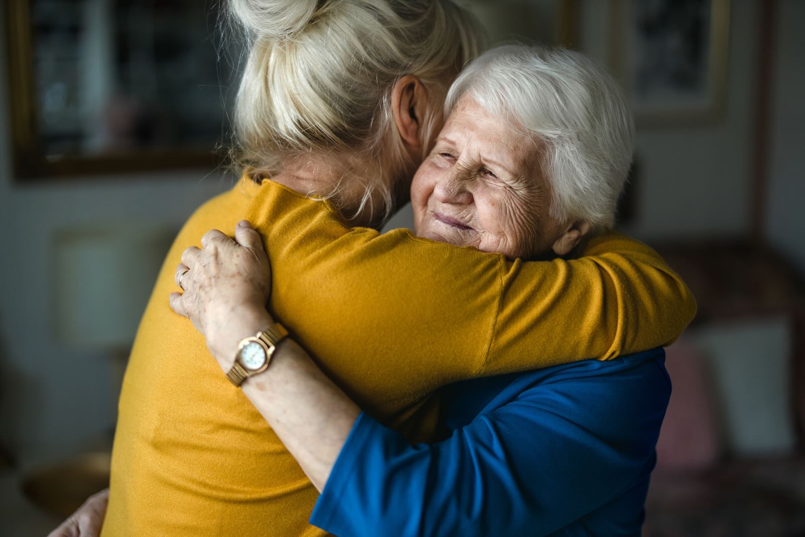 Young woman in a yellow shirt hugging an elderly woman wearing jewelry