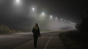 Woman walks on the sidewalk on one side of the road at night, streetlamp visible overhead.