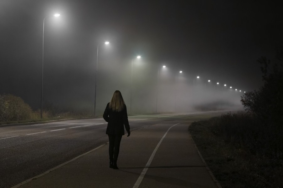 Woman walks on the sidewalk on one side of the road at night, streetlamp visible overhead.