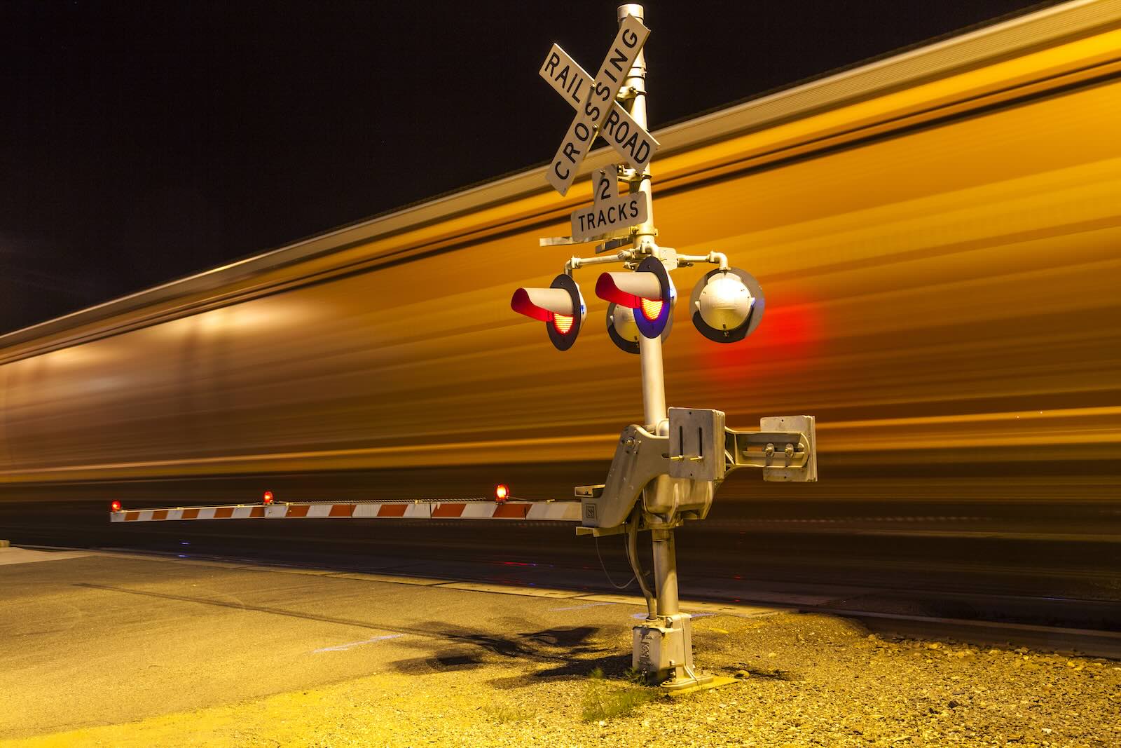 The blur of a train on the tracks crossing the road at night.