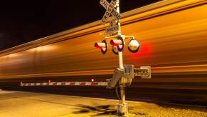 The blur of a train on the tracks crossing the road at night.
