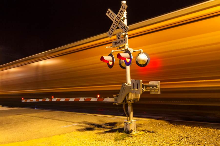 The blur of a train on the tracks crossing the road at night.