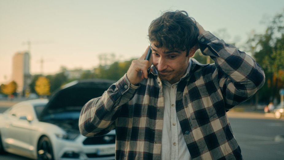 A driver uses his cell phone in the foreground with a broken down car in the background