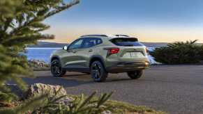 A sage green Chevrolet Trax compact SUV parked in left rear angle view near water with an evergreen tree branch in the left foreground