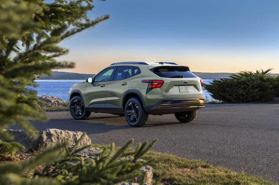 A sage green Chevrolet Trax compact SUV parked in left rear angle view near water with an evergreen tree branch in the left foreground