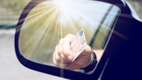A man's hand holding a driver's license reflected in a car's side mirror