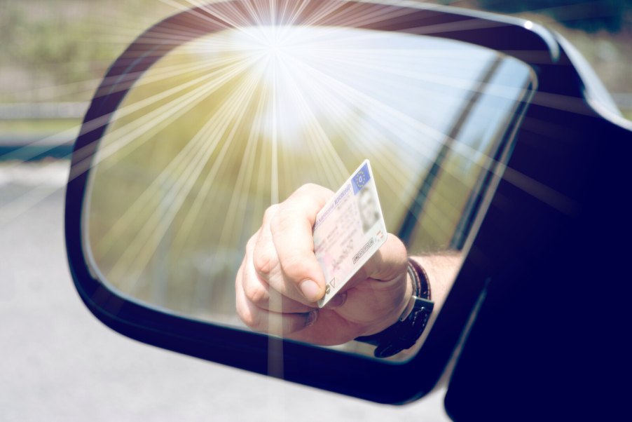 A man's hand holding a driver's license reflected in a car's side mirror