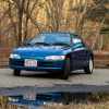 Blue Honda Beat convertible parked in front of the fall woods in Massachusetts, its reflection visible in a puddle in the foreground.