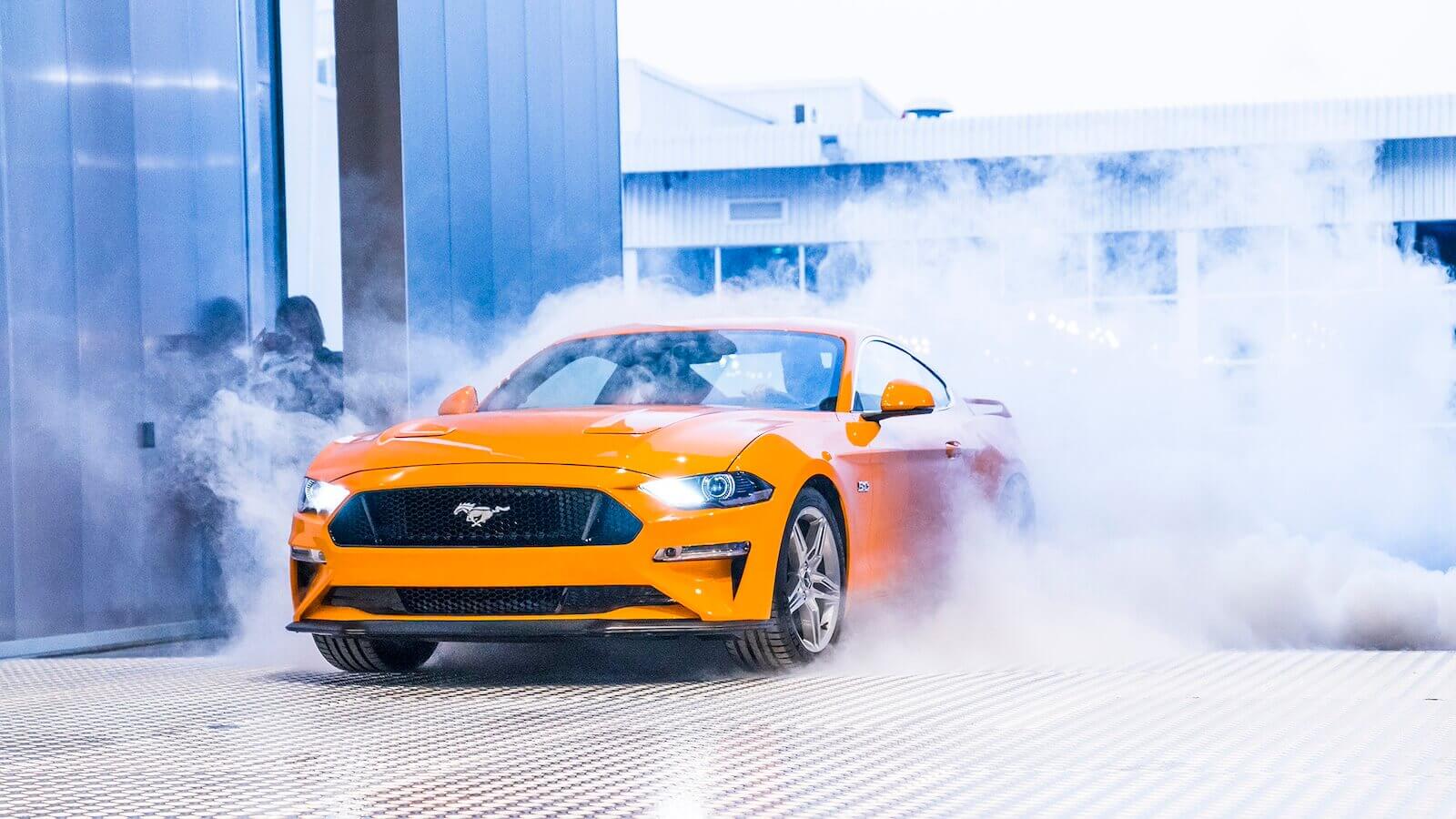 A bright-orange 2018 Ford Mustang GT kicks up smoke from its rear tires.