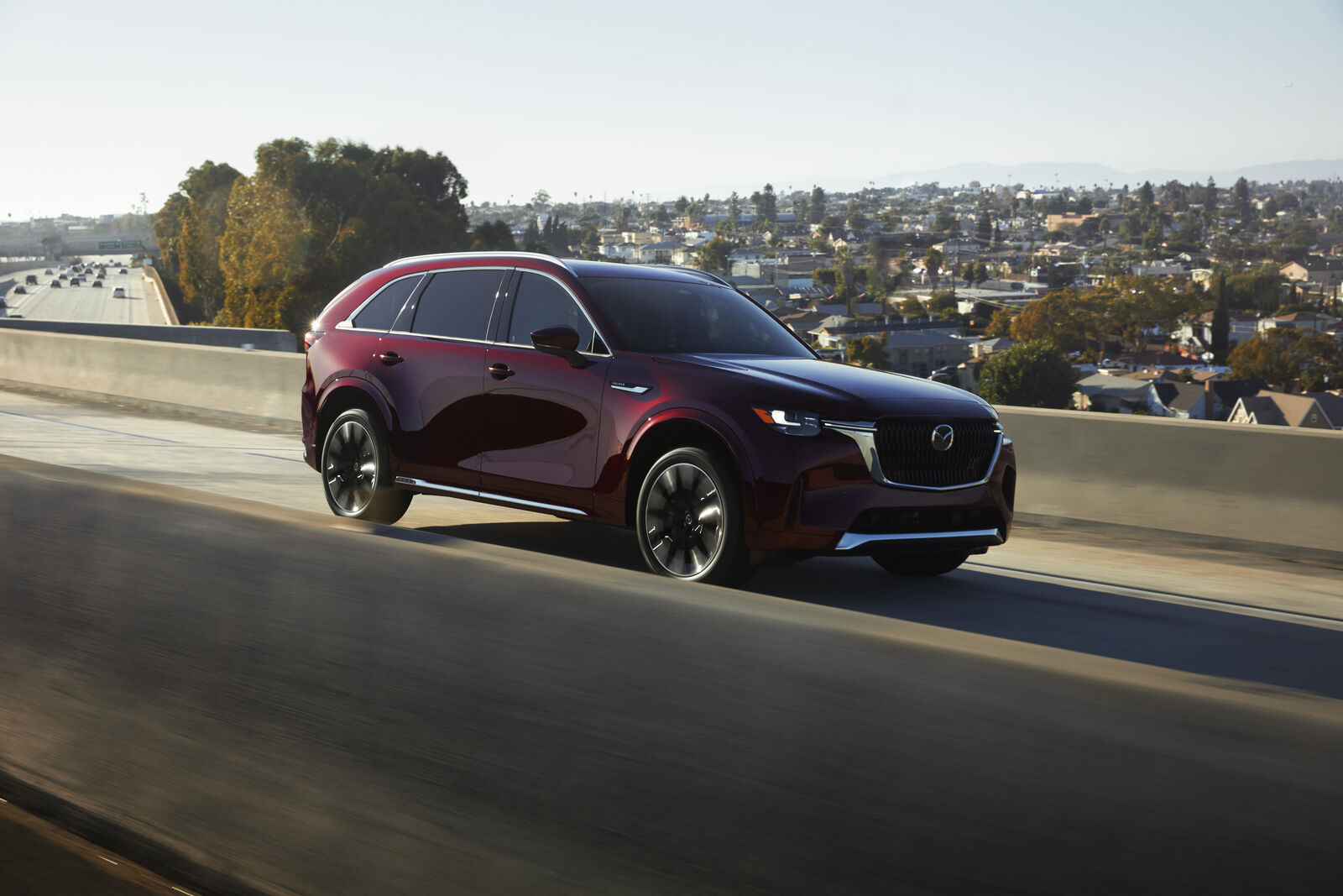 A burgundy metallic 2024 Mazda CX-90 SUV driving on the highway in right side angle view