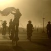 Burning man festival goers travel through the festival on bicycles and stilts during a dust storm, only their silhouettes visible.