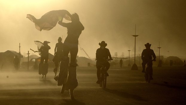 Burning man festival goers travel through the festival on bicycles and stilts during a dust storm, only their silhouettes visible.