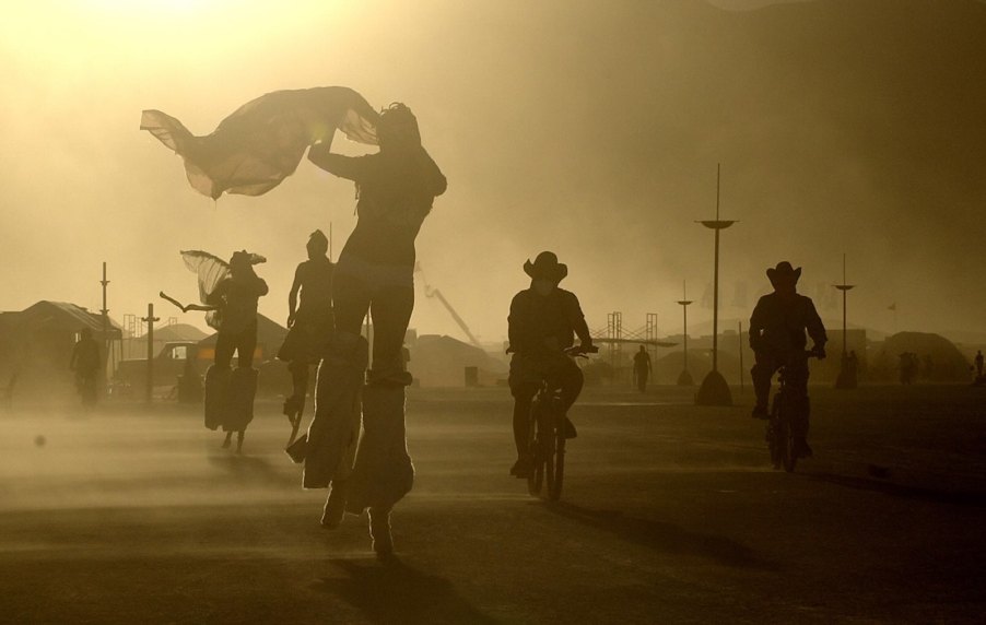 Burning man festival goers travel through the festival on bicycles and stilts during a dust storm, only their silhouettes visible.