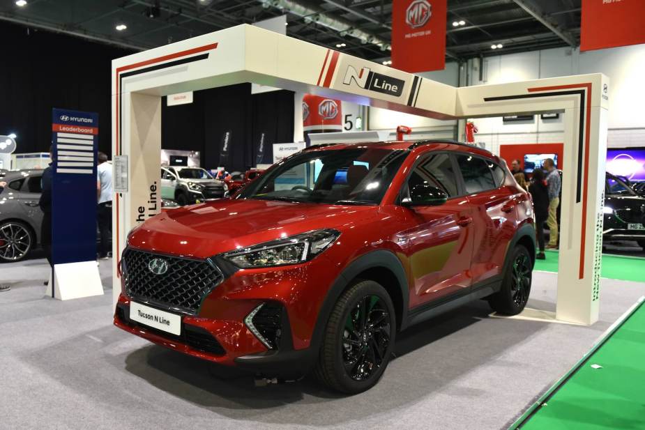 Red Hyundai Tucson N Line crossover SUV at an auto show, a display in the background.