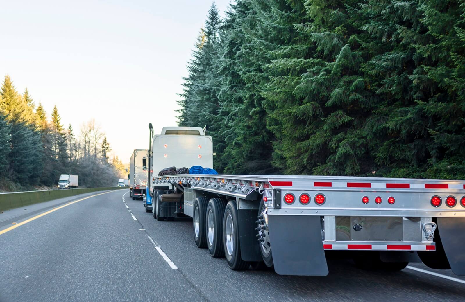 Semi truck flatbed trailer with a Mansfield bar bumper hanging down beneath its bed, driving along a highway.