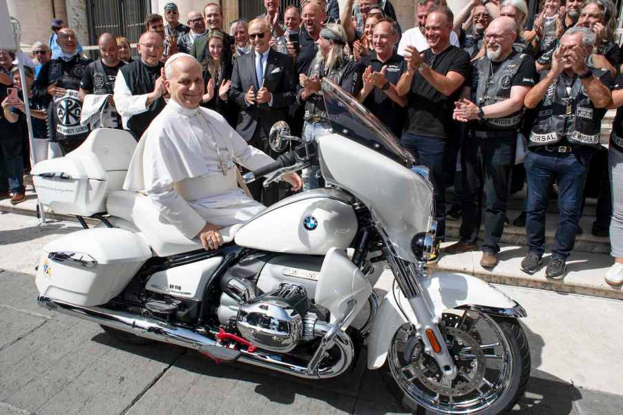 Pope Leo XIV sits on a BMW cruiser motorcycle, while the Jesus Bikers club stands behind him and cheers.