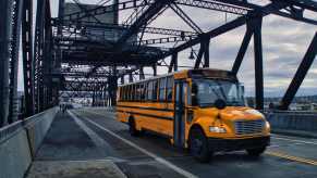 Yellow school bus drives over a steel bridge, a dark sky visible overhead.