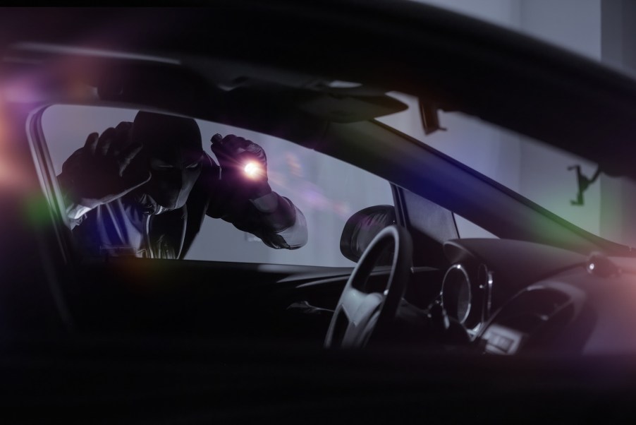 Masked thief with a flashlight looks inside a car after dark.