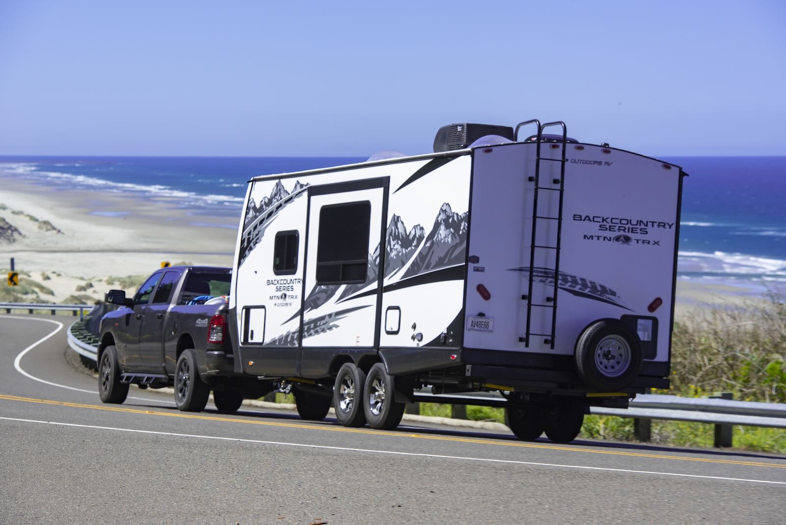 White camper trailer hooked to a black pickup truck by the roadside, the beach and ocean visible in the background.