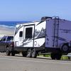 White camper trailer hooked to a black pickup truck by the roadside, the beach and ocean visible in the background.