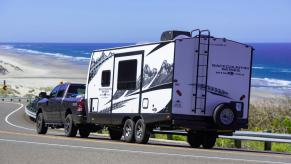 White camper trailer hooked to a black pickup truck by the roadside, the beach and ocean visible in the background.