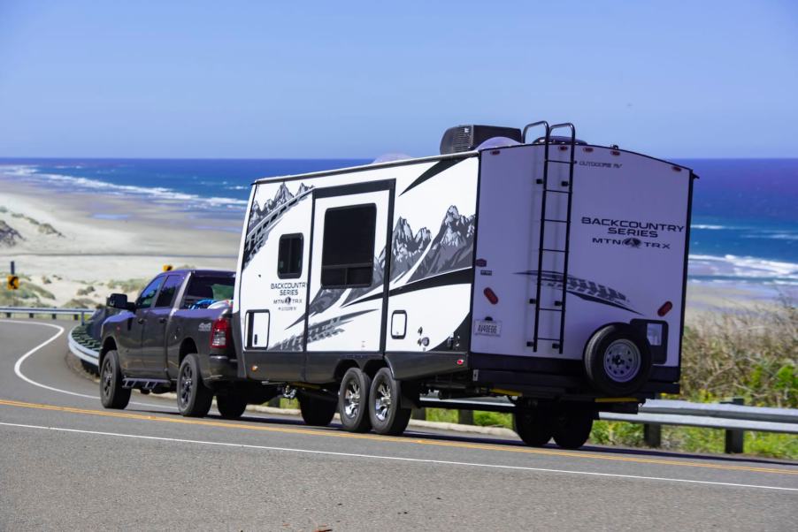 White camper trailer hooked to a black pickup truck by the roadside, the beach and ocean visible in the background.