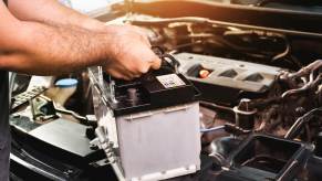 A mechanic's arms and hands holding a car battery above an engine bay