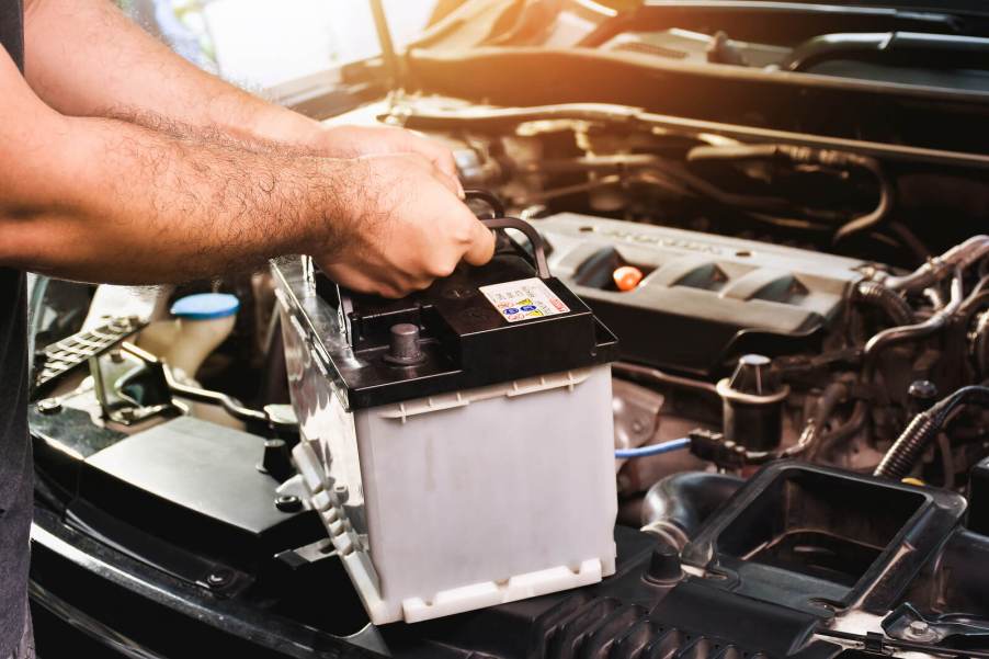 A mechanic's arms and hands holding a car battery above an engine bay
