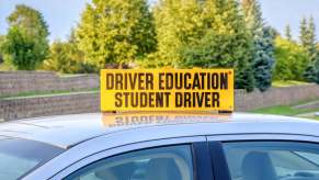 A yellow sign on a car roof reads "DRIVER EDUCATION STUDENT DRIVER"