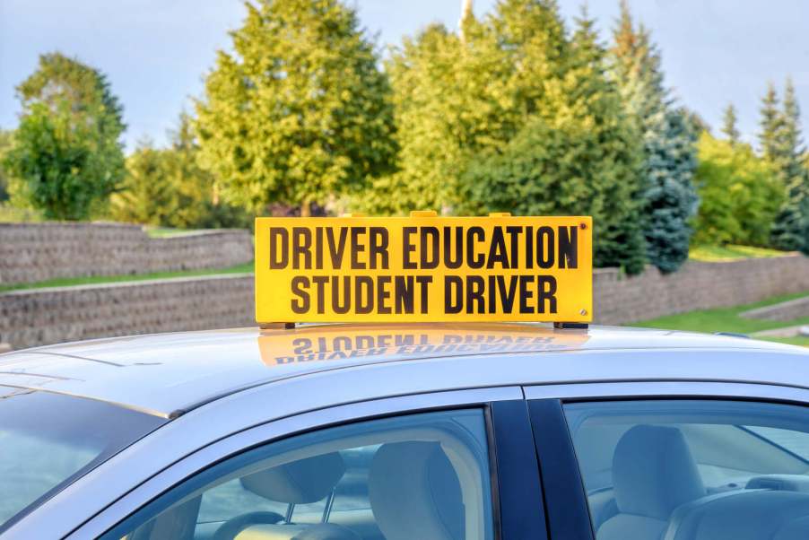 A yellow sign on a car roof reads "DRIVER EDUCATION STUDENT DRIVER"