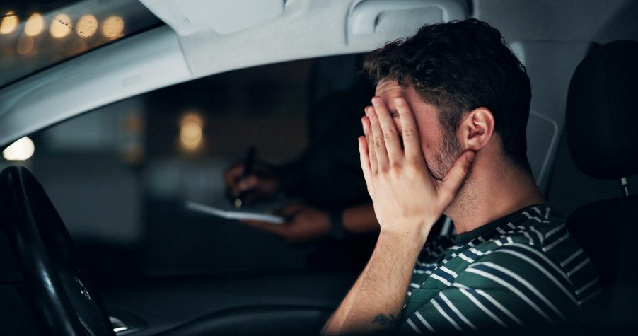 A driver covers their face with a police officer standing at opposite window at night in a depiction of a DUI investigation